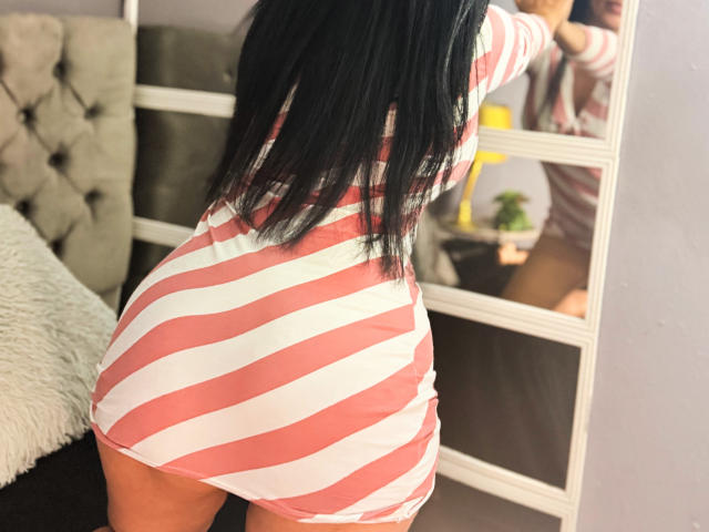 VeraCruz wearing a pink and white striped dress, butt facing mirror, showing her curvy ass and long black hair, reflecting her upper body in the mirror.