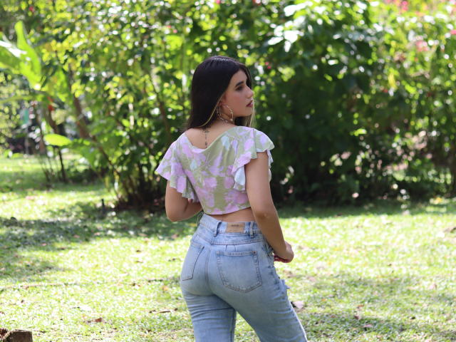 KatieDesirX with long dark hair, pink floral crop top, and high-waisted blue jeans, showing her ass, standing in a green grassy area, looking over her shoulder.