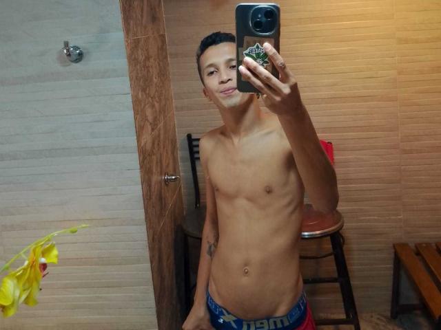 NickHotX, senza camicia, che tiene in mano un telefono, selfie allo specchio, capelli neri corti, pelle marrone chiaro, capezzoli piccoli, magro, indossa biancheria intima blu con la scritta "BENJEN" sull'elastico, uno sgabello di legno sullo sfondo..