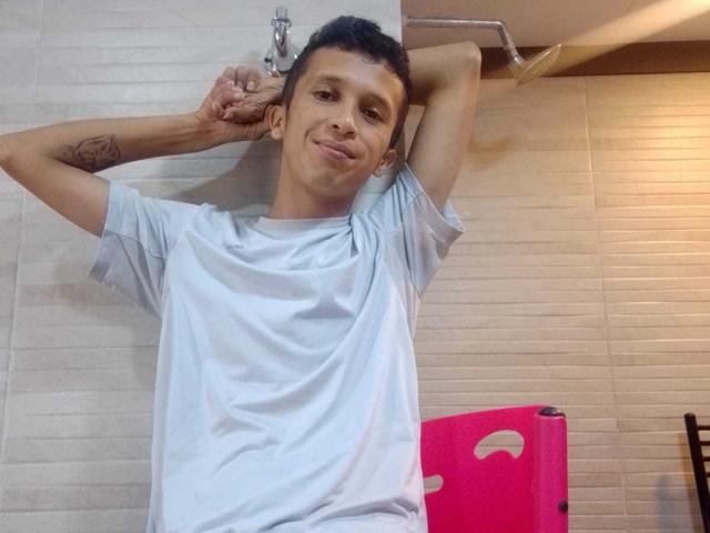 NickHotX, con cabello negro corto, con los brazos detrás de la cabeza, vistiendo una camiseta blanca, sonriendo ligeramente, tiene un tatuaje en el brazo derecho, está de pie en un baño con azulejos y un inodoro rosa..