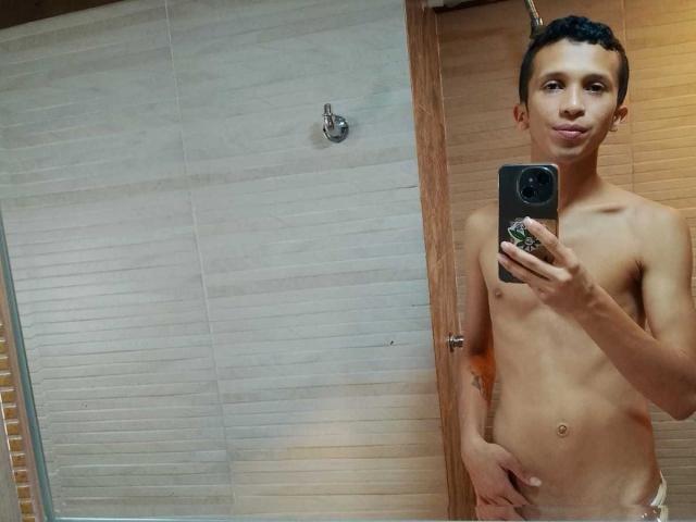 Nick, con indosso solo un asciugamano, è magro, con capelli corti e scuri, un pene piccolo e flaccido, tiene in mano un telefono, fa una smorfia, sullo sfondo si vede una porta di legno, è in piedi nel bagno, si sta facendo un selfie allo specchio..