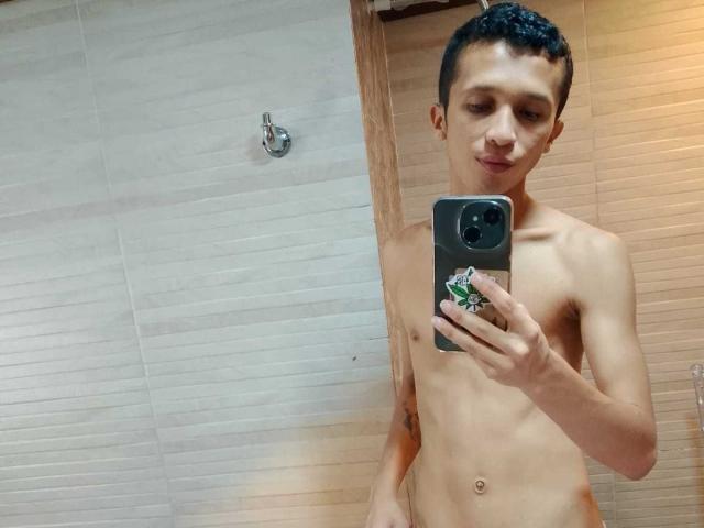 NickHotX, slank met kleine borsten, kort zwart haar, houdt een telefoon vast, maakt een spiegelzelfportret, naakt, bruine huid, kleine tatoeage op de linkerarm, kijkt naar beneden, staat in de badkamer..
