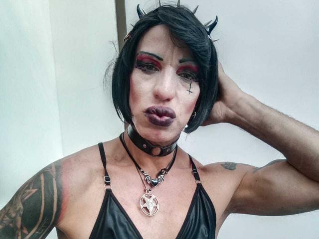LilithMadame with black horns, dark lipstick, red eyeshadow, black choker, leather top, tattooed arm, and pentagram necklace, looking at camera with hand in hair.