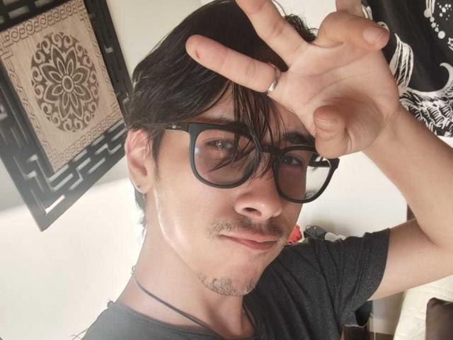 KingDanny met een bril, zwart haar, een snor en een ring aan de wijsvinger, die een vrede-teken maakt, gekleed in een zwart shirt, met een ketting en een lichte grijns, op een selfie..