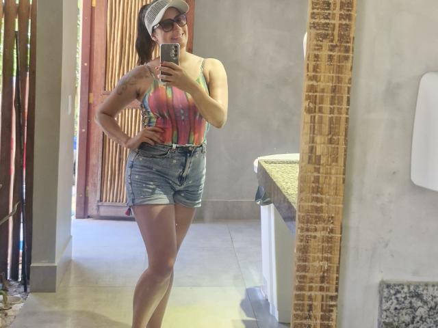A mirror selfie of DarkAng, a woman with light brown skin and dark brown hair in a ponytail, wearing a white cap, dark sunglasses, a colorful striped tank top, and denim shorts. She stands with one hand on her hip, holding a smartphone in the other. She has a toned physique with visible muscle definition in her legs. The background includes a wooden door and a granite countertop. The image has a casual, everyday style.