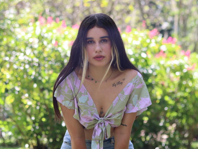 KatieDesirX, with long dark hair and blonde highlights, leans forward in a pink and green floral top, showing her tits and tattoos, against a green, sunlit background.
