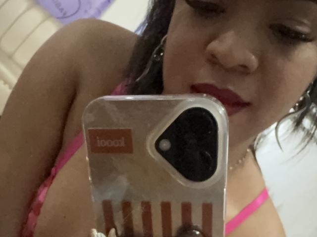 AliceBooty taking a selfie with a transparent phone case, showing her tits in a pink bra, red lips, and short dark hair, holding the phone with her right hand.