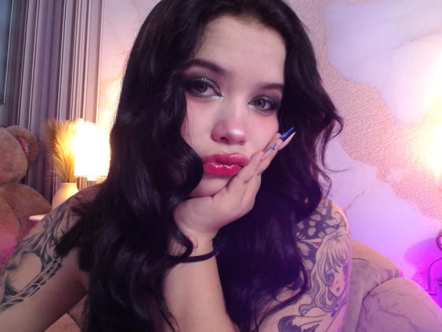 AryButty with long black hair, red lipstick, and blue nails, resting her face on her hand, showing tattooed arms, looking seductively at the camera.