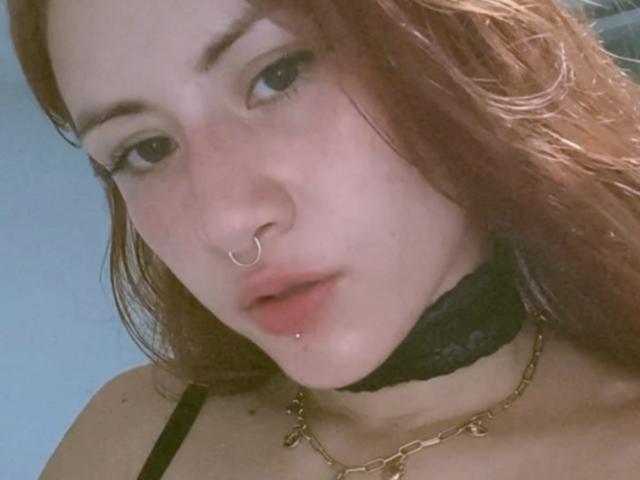 MalenaJones with pale skin, red hair, nose ring, lip piercing, black lace choker, gold chain, black strap, slightly parted red lips, looking at camera, soft light.
