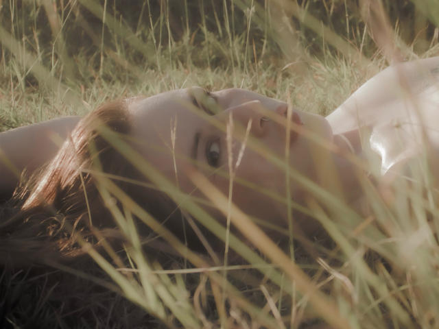 MalenaJones lies on her back in tall grass, her face partially obscured by the blades. Sunlight casts a soft glow on her fair skin. She has brown hair that spreads out around her head. Her eyes are wide and looking slightly upward. The grass is green and yellow, with some blades in the foreground. The image has a natural, slightly blurred look, emphasizing the grass and MalenaJones' face. The composition focuses on the contrast between her skin and the grass.