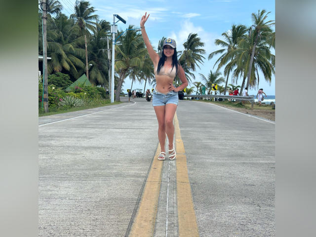 GraceClarke with long dark hair, wearing a white cap, beige bra, and blue denim shorts, standing on a road, waving with one hand, showing her ass in white sandals.
