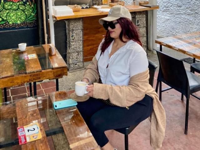ElyRoss, with big ass and large tits, sits at a wooden table, wearing a beige hat, sunglasses, white shirt, and black pants, holding a white cup, on a patio.