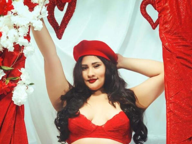 ElyRoss with long black hair, red beret, and red lingerie, arms raised, smiling, showing her tits, surrounded by red and white floral decorations.