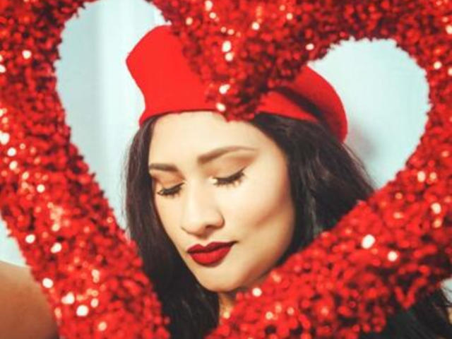 ElyRoss with red sequined heart frame, wearing red hat and lipstick, dark hair, and closed eyes, showing off her red-lipped, cute face. Boobs partially visible, looking down.