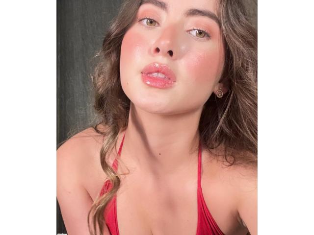 JadeCarter has shiny pink lips, a slight tan, and wavy brown hair, wearing a red halter top, with a cute, pouty expression, and her tits barely visible.
