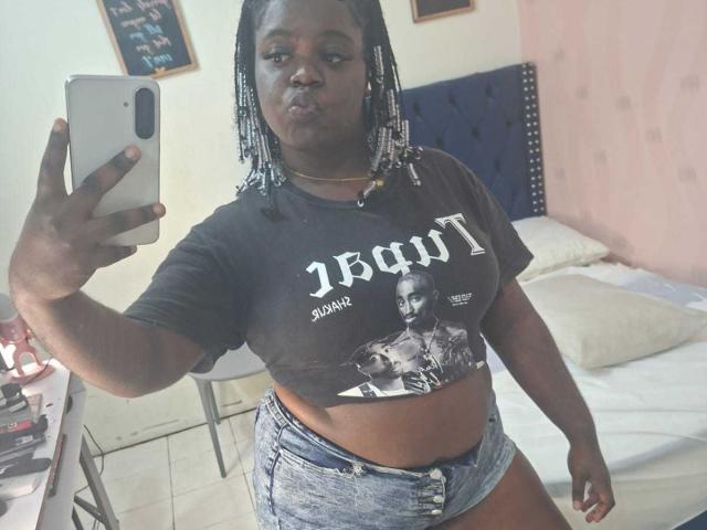 LikeBlackNuts with dark skin, braided hair, wearing a black crop top with a photo of a man and denim shorts, taking a selfie in a bedroom, holding a white phone.