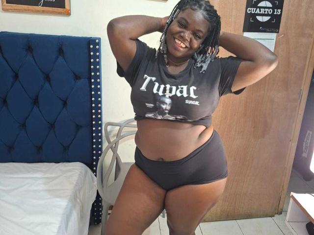 LikeBlackNuts mit dunkler Haut, geflochtenen Haaren, lächelnd, trägt ein schwarzes Tupac-Shirt und schwarze Unterwäsche, Hände hinter dem Kopf, steht in einem Schlafzimmer mit einem blauen, gepolsterten Kopfteil..