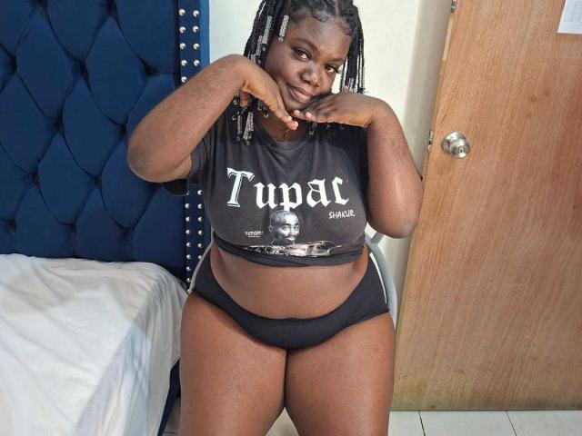 LikeBlackNuts, a dark-skinned woman with braided hair, showing her tits in a black Tupac shirt and black panties, smiling with hands near her face.