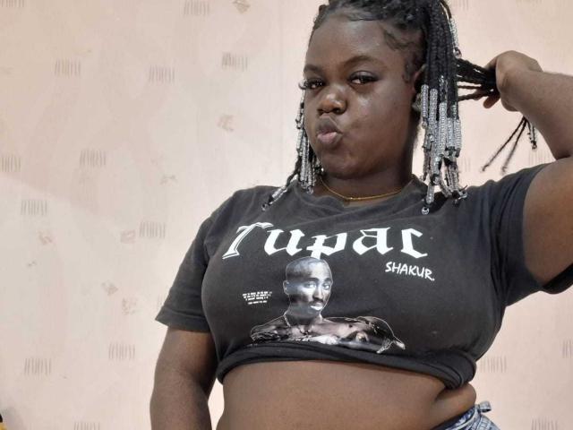 LikeBlackNuts, una mujer de piel oscura con trenzas, que lleva una camiseta negra de Tupac Shakur, tiene el ceño fruncido, una mano en el pelo, y muestra sus pechos y un poco de su abdomen..