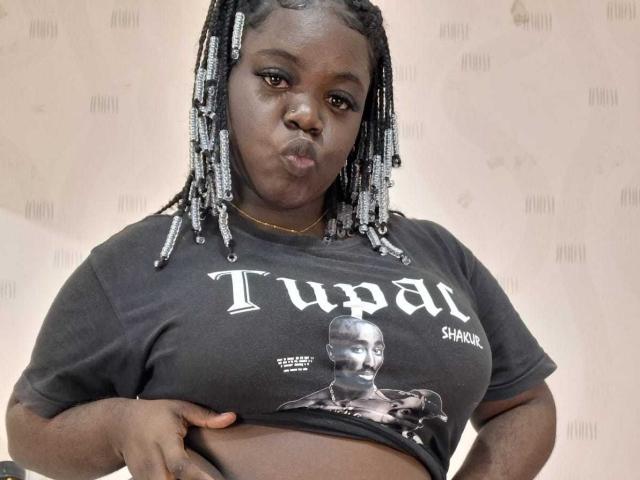 Net als BlackNuts, met donkere huid en gevlochten haar, rolt ze haar lippen op, draagt een zwart T-shirt met een afbeelding erop, tilt haar shirt op om haar buik te laten zien, heeft een gouden ketting, en de achtergrond is effen..