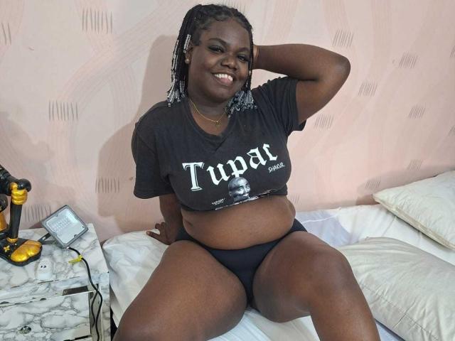 CommeLikeBlackNuts, femme de couleur, corpulente, avec des cheveux tressés, portant un t-shirt noir et des sous-vêtements, assise sur un lit, souriante, une main dans les cheveux, ventre visible, oreillers blancs, lampe jaune..