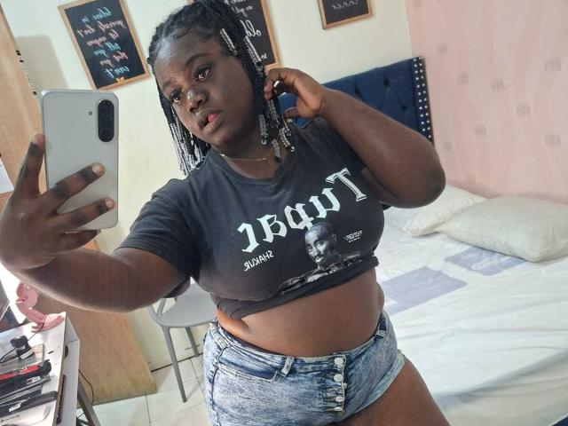 LikeBlackNuts, met donkere huid, gevlochten haar, een zwart T-shirt dat haar borsten laat zien, en blauwe denim shorts, die een selfie maakt in een slaapkamer met een bed..