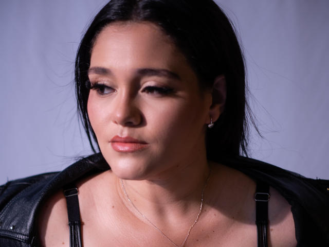 Astrid Williams, avec ses cheveux foncés, portant une veste en cuir noir, a un maquillage discret et une expression légèrement sérieuse, et on peut apercevoir ses seins, qui semblent doux, ainsi qu'un collier délicat..