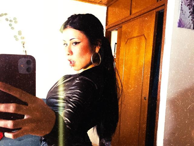 GaiaJoness with black hair, wearing a shiny black leather top, looking back at a mirror, holding a phone, large hoop earrings, slightly pouted lips, wooden door in background.