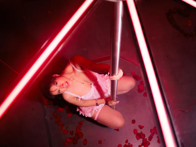 HanaJoud kneels by a pole, wearing pink lingerie and fishnet stockings, with red neon lights and rose petals on the floor. Her tits are visible, and she's holding the pole.