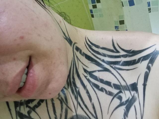 MillaSouReety's upper chest with black tribal tattoos, showing part of her mouth and chin. Her lips are slightly parted, and she has some acne on her skin.