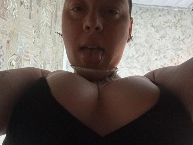 MillaSouReety with tongue out, looking down at her large boobs in a black top, wearing a choker, short dark hair, and small cross earrings, in a low-light selfie.