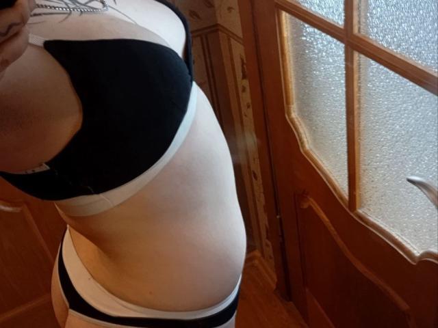 MillaSouReety's side profile showing black and white bra and panties, with her tits in a black bra and her ass slightly visible, standing near a wooden door.