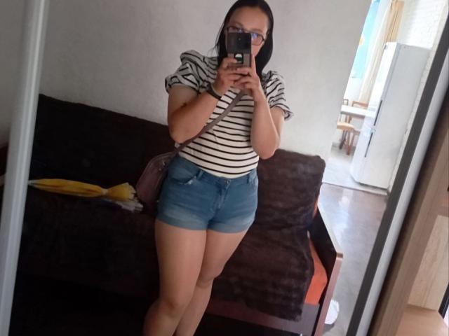MillaSouReety stands in front of a mirror, wearing a striped shirt and blue denim shorts, showing her ass and holding a phone, with glasses and dark hair.