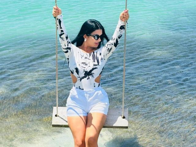 XimenaBlaze with black hair, sunglasses, white and black long-sleeve top, cutout sides, white shorts, sitting on a wooden swing in clear water, holding ropes.