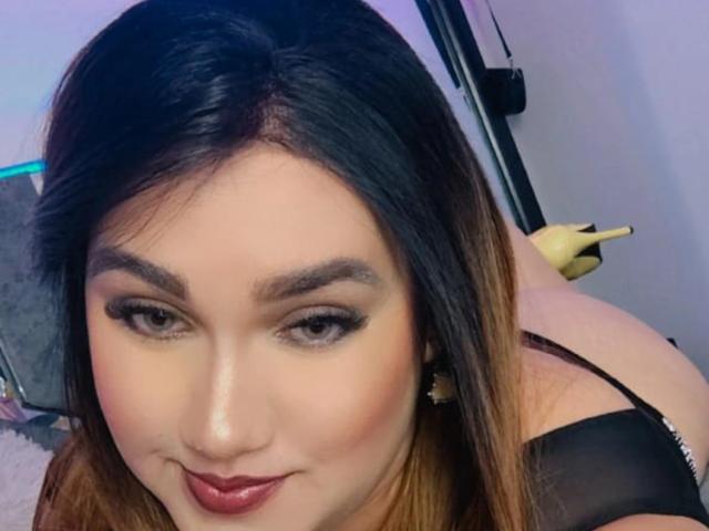 StaceyLatinHot with dark brown hair, thick eyebrows, black eyeliner, and red lipstick, wearing a black top, looking at the camera with a slight smile, showing her ass.