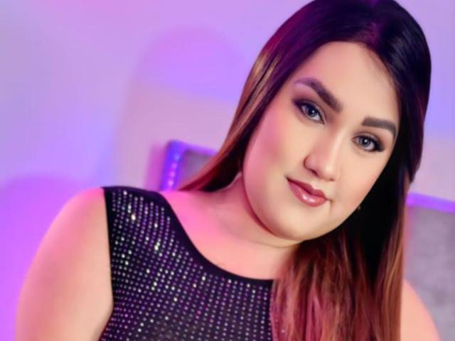 StaceyLatinHot has long brown hair, a cute face with full lips, and is wearing a sparkly black dress. Her eyes are blue and she's smiling softly.