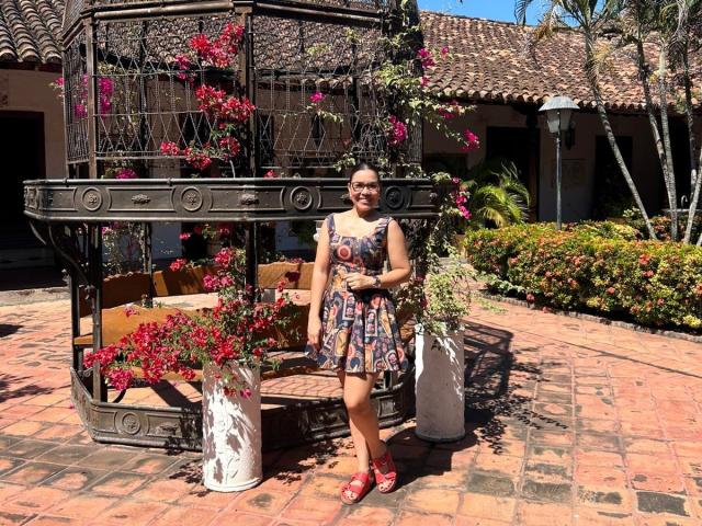 AnnaJaimes stands in a sunlit courtyard wearing a colorful, patterned dress with red sandals, in front of a metal birdcage with blooming pink flowers, holding a white vase.