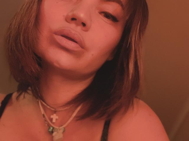 ElizavetaCream with pouty lips, short brown hair, and a cross necklace, wearing a black top, under warm lighting. Her nose is slightly upturned.
