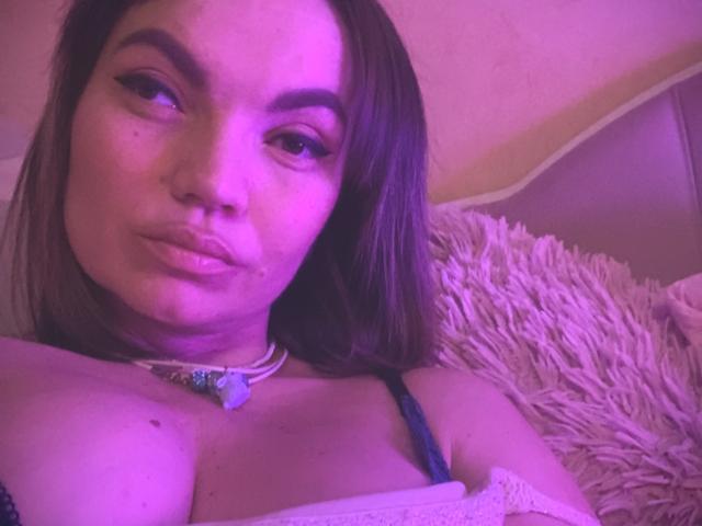 ElizavetaCream with purple-lit face, pouty lips, and brown hair, wearing a necklace with a blue gem, showing her tits in a white top, lying on a fluffy pillow.
