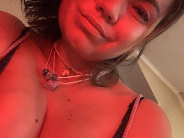 ElizavetaCream with red-lit skin, brown wavy hair, smiling slightly, wearing a pink-strapped top, necklace with purple crystal and cross, showing her tits.