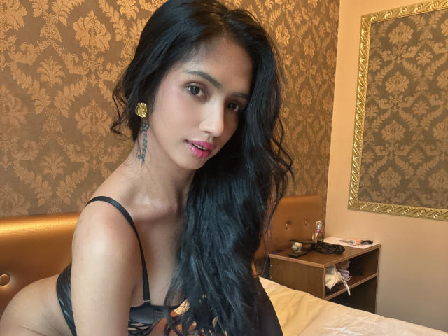 DannaWalton with long black hair, wearing black lingerie, showing her tits and pink lipstick, sitting on a bed, looking at the camera, gold earrings, and a tattoo on her neck.