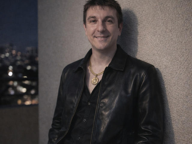KEV69, sonriendo, vistiendo una chaqueta de cuero negro sobre una camisa negra, una cadena de oro con un colgante circular, apoyado contra una pared texturizada, noche, luces de la ciudad en el fondo..