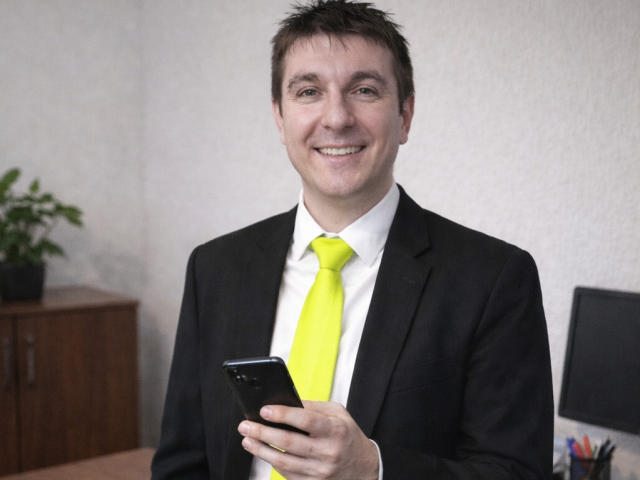 KEV69 with short brown hair, smiling, wearing a black suit, white shirt, and bright yellow tie, holding a black smartphone, standing in a room.