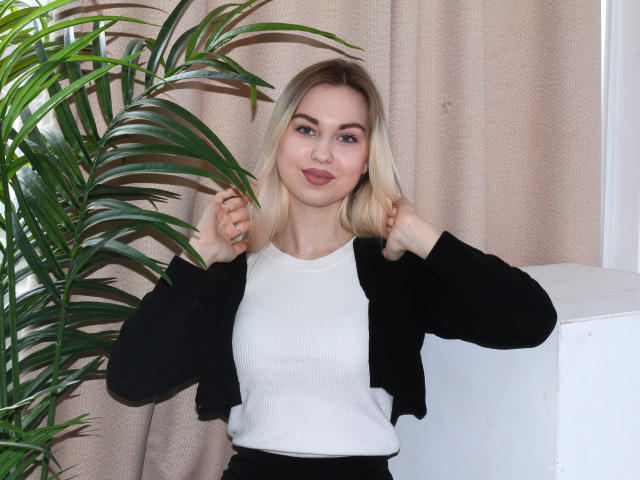 NicoleHrum with blonde hair, white ribbed top, black cardigan, holding hair, green plant on left, beige curtain background, standing, smiling, showing small ass, cute, attractive.