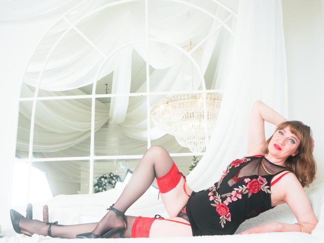 SquirtyCumShow lies on a white bed in a brightly lit room. She wears a black mesh lingerie dress with red rose floral patterns, red garter straps, and black stockings with red tops. She has red lipstick, light skin, and brown hair. Her right arm is raised, and her left arm rests on her head. She wears black high heels. A large, ornate chandelier hangs in the background. The image has a bright, almost overexposed look, emphasizing the white bed and sheer curtains.