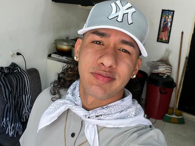 DamiamPhill, con i capelli castano ricci, indossa un berretto grigio dei New York Yankees, una bandana bianca e orecchini dorati, ha un'espressione neutra ed è vestito con una maglietta grigia, che rivela una leggera abbronzatura..