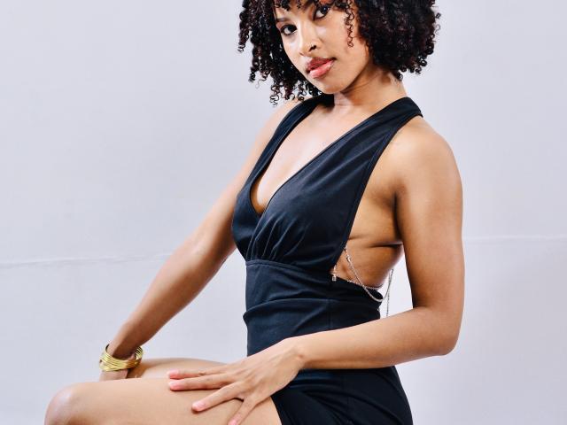 Plaissir69 with curly black hair, wearing a black halter dress that exposes her side and shows her tits, sitting with a gold bracelet on her wrist.