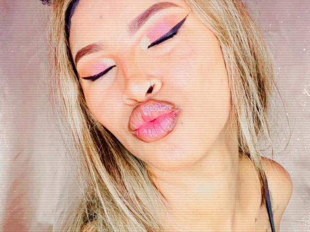 Plaissir69's face, with pink eyelids and black eyeliner, has eyes closed and pink, glossy lips puckered. Blonde hair, fair skin, and a black strap are visible.