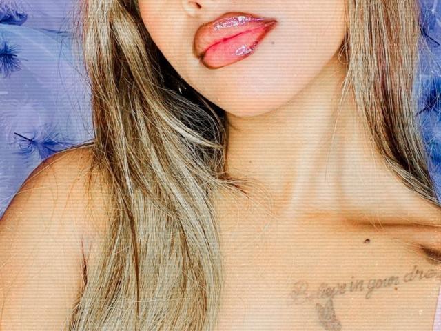 Plaissir69's glossy pink lips are slightly open, tongue out, with long blonde hair falling over her chest. Her bare shoulders are visible, showing a small mole.