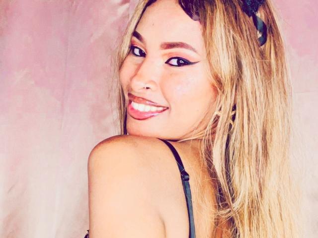 Plaissir69 with long blonde hair, black eyeliner, and a smile showing pink lipstick, looking over her shoulder, wearing a black strap. Her tits are partially visible.